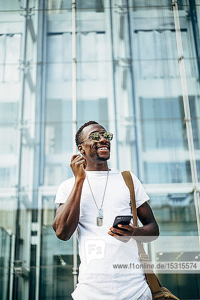 Glücklicher junger Mann mit Handy in der Stadt  der Kopfhörer einsteckt