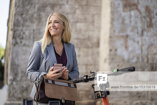 Porträt einer lächelnden blonden Geschäftsfrau mit Aktentasche  Mobiltelefon und Elektroroller