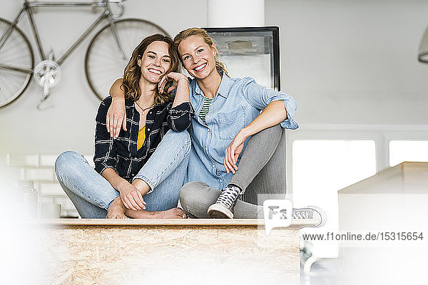 Two young women sitting cross-legged on the counter of their coffee shop with arms aroug