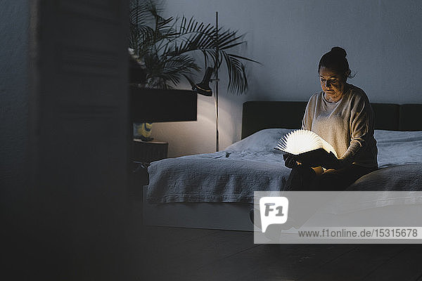 Senior woman sitting on bed holding glowing book