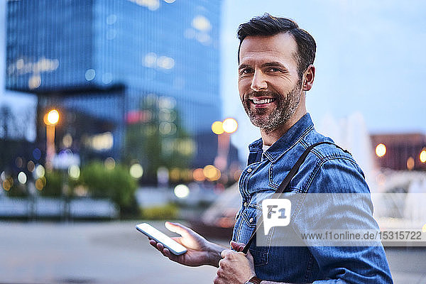 Porträt eines lächelnden Mannes  der abends in der Stadt steht und ein Smartphone benutzt