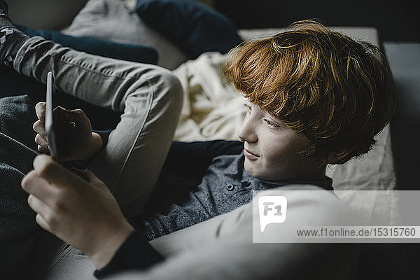 Rothaariger Junge liegt mit digitalem Tablett auf der Couch