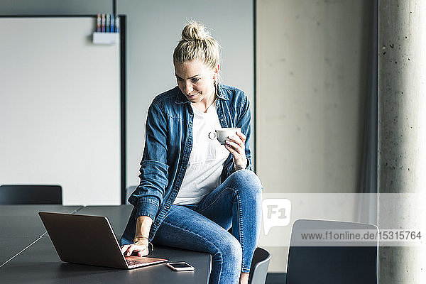 Businesswoman with cup of coffee sitting on table in office using laptop