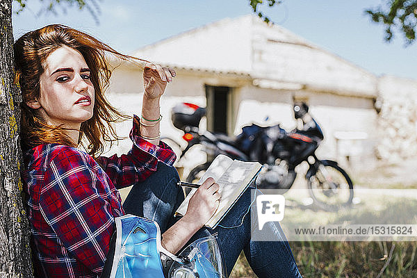 Portrait of redheaded motorcyclist leaning against tree trunk taking notes  Andalusia  Spain