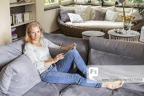 Porträt einer blonden reifen Frau  die sich zu Hause auf der Couch mit einem digitalen Tablet entspannt