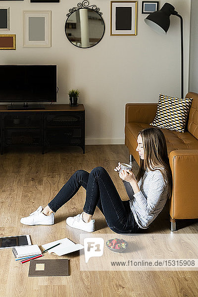 Entspannte junge Frau sitzt zu Hause auf dem Boden und trinkt Kaffee