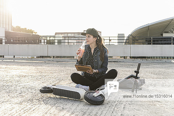 Junge Frau mit Roller  Getränk und Tablette sitzt bei Sonnenuntergang auf dem Parkdeck