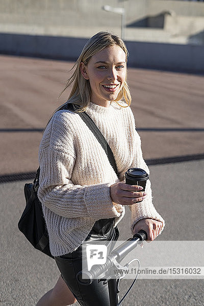 Porträt einer lächelnden jungen Frau mit Sporttasche  Kickroller und Kaffee zum Mitnehmen
