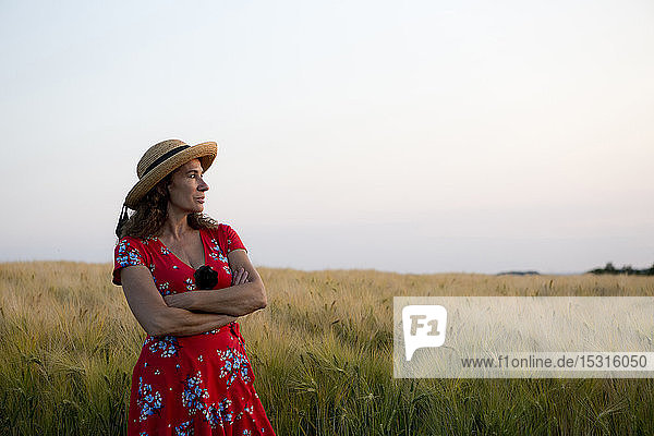 Woman wearing straw hat and red summer dress with floral design standing in front of grain field