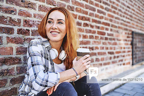 Young woman sitting against brick wall holding cup of coffee and looking away