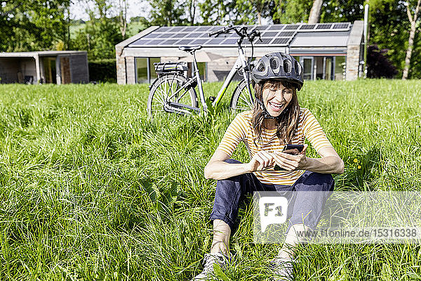 Lachende Frau mit Fahrrad mit Handy auf einer Wiese vor einem Haus