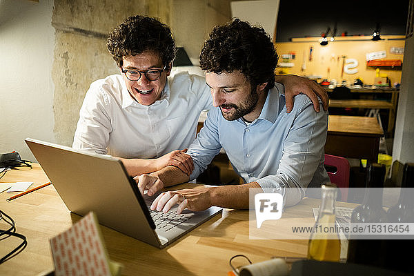 Zwei glückliche Kollegen benutzen gemeinsam einen Laptop in der Dunkelheit