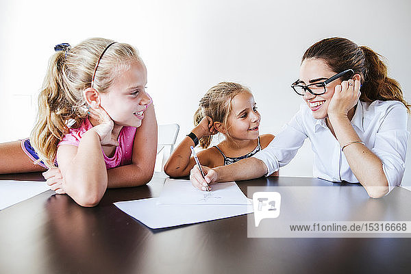 Glückliche Lehrerin sitzt am Schreibtisch mit zwei Schulmädchen  die auf Papier schreiben