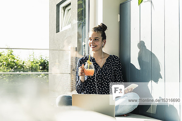 Lächelnde junge Frau mit Laptop  Handy und gesundem Getränk zu Hause