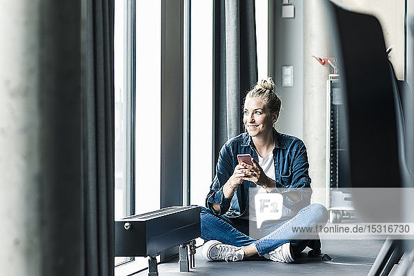 Lächelnde Geschäftsfrau sitzt im Büro mit Handy auf dem Boden