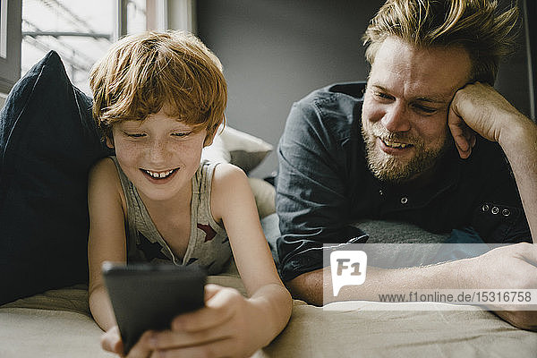 Porträt von glücklichem Vater und Sohn  die zusammen auf der Couch liegen und auf ihr Handy schauen