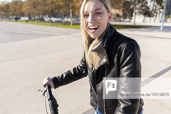 Porträt einer lachenden jungen Frau mit Kickroller in schwarzer Lederjacke