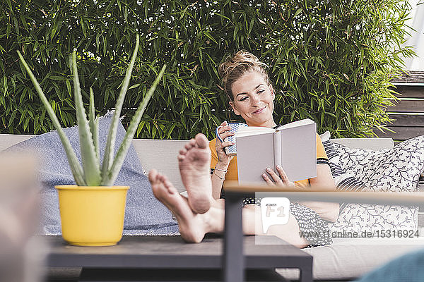 Entspannte Frau sitzt zu Hause auf einer Couch auf der Terrasse und liest ein Buch