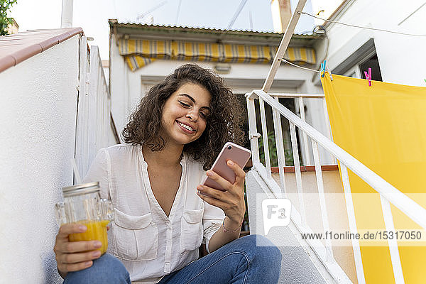 Lächelnde junge Frau auf der Treppe sitzend mit Zelle und Orangensaft