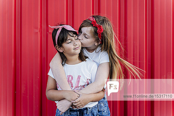 Zwei Schwestern stehen vor einer roten Wand und küssen sich