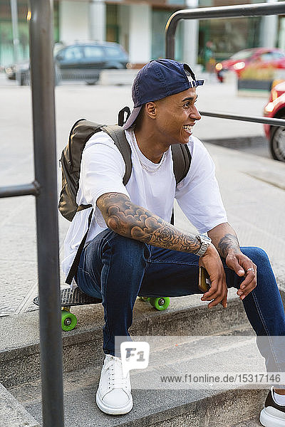 Tätowierter junger Mann mit Rucksack und Smartphone sitzt auf einem Skateboard und schaut in die Ferne