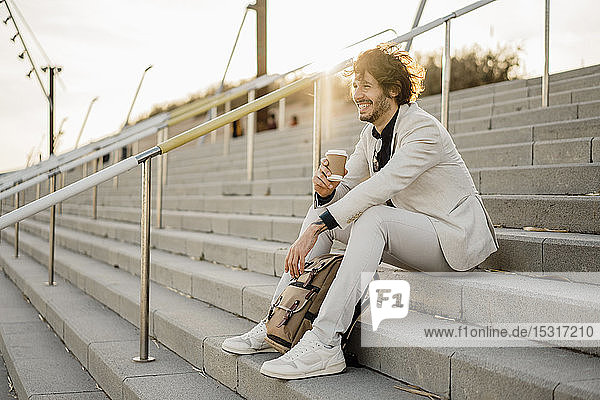 Lachender Geschäftsmann mit Kaffee zum Mitnehmen und Rucksack auf der Treppe im Freien sitzend