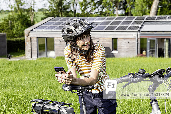 Lächelnde Frau mit Fahrrad und Handy auf einer Wiese vor einem Haus