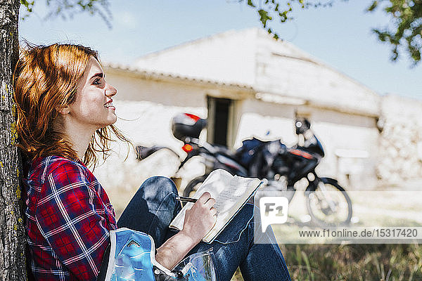 Smiling motorcyclist leaning against tree trunk taking notes  Andalusia  Spain