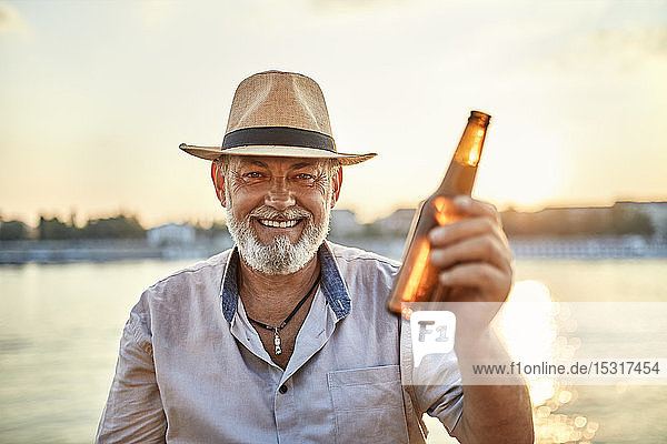 Porträt eines glücklichen älteren Mannes  der bei Sonnenuntergang am Flussufer ein Bier trinkt