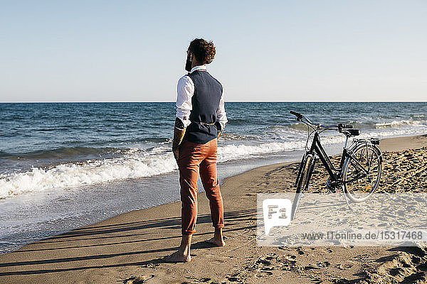Gut gekleideter Mann mit seinem Fahrrad am Strand stehend