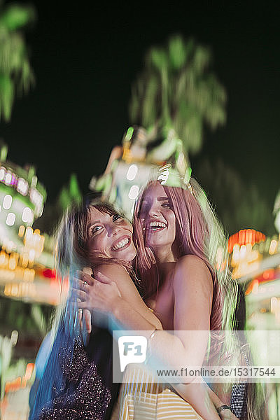 Porträt von zwei aufgeregten jungen Frauen  die sich nachts auf einem Jahrmarkt umarmen