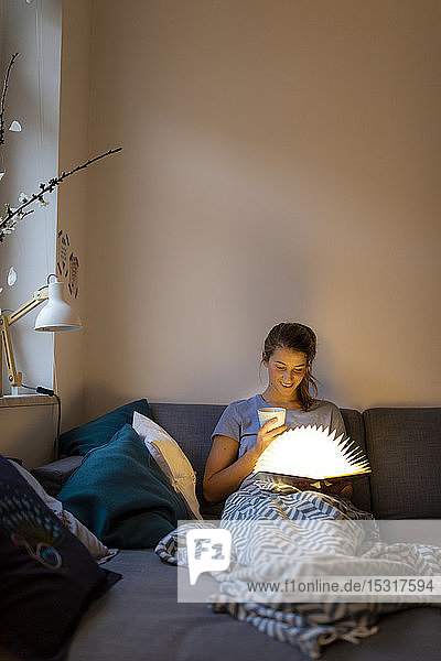 Junge Frau liest zu Hause auf dem Sofa ein illuminiertes Buch