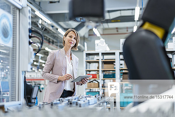 Geschäftsfrau mit Tablette in einer modernen Fabrikhalle mit Blick auf Roboter