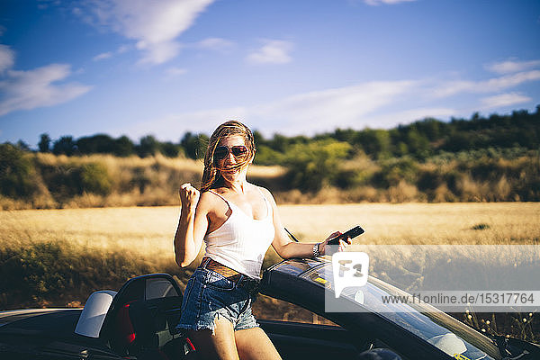 Woman standing in her convertible  holding smartphone  happy about good news
