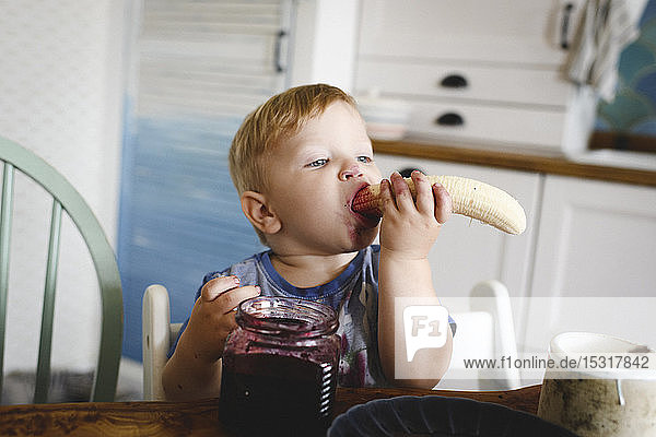 Cute little boy eating banana with blueberry jam in the kitchen