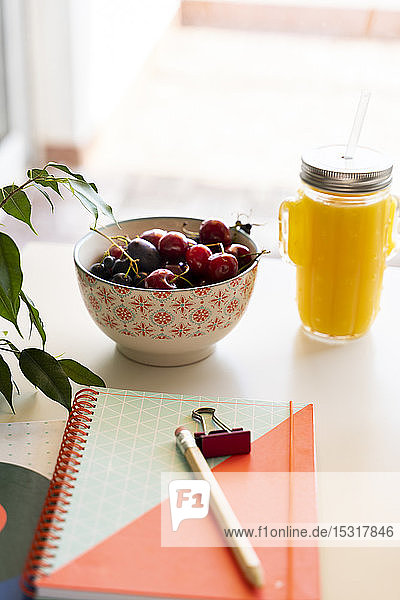 Orangen-Smoothie und Kirschschale auf Tisch mit Notizbuch und Bleistift