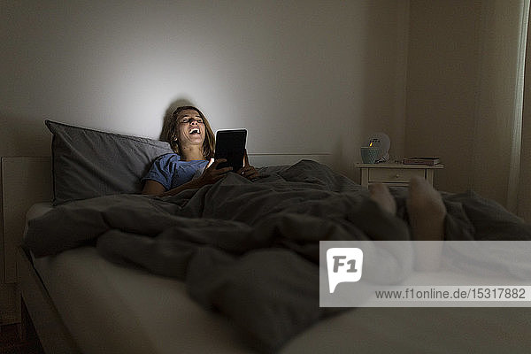 Laughing young woman lying in bed at home at night using tablet