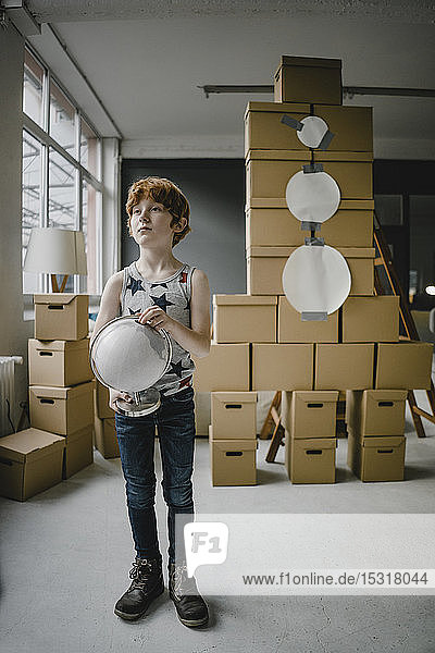 Porträt eines rothaarigen Jungen mit Globus  der vor einer aus Pappkartons gebauten Rakete steht
