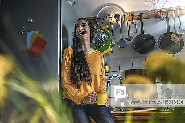 Lachende junge Frau sitzt zu Hause auf dem Küchentisch