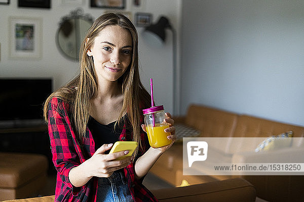 Porträt einer lächelnden jungen Frau mit Mobiltelefon und gesundem Getränk zu Hause