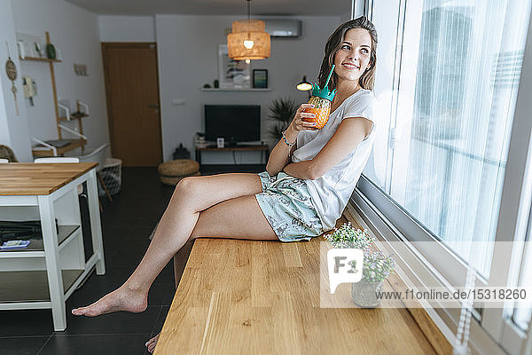 Lächelnde junge Frau sitzt mit einem Getränk auf der Küchentheke und schaut aus dem Fenster
