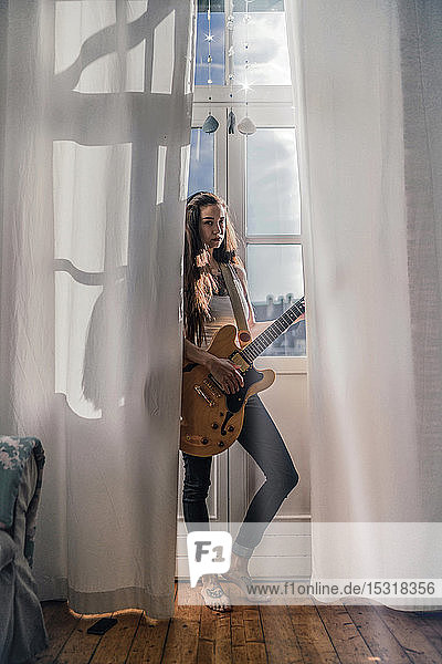 Seriöse junge Frau  die zu Hause mit einer Gitarre am Fenster steht
