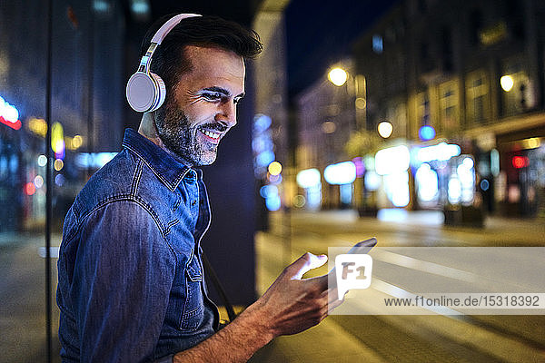 Lächelnder Mann mit Kopfhörern  der ein Smartphone benutzt  während er in der Stadt auf den Nachtbus wartet