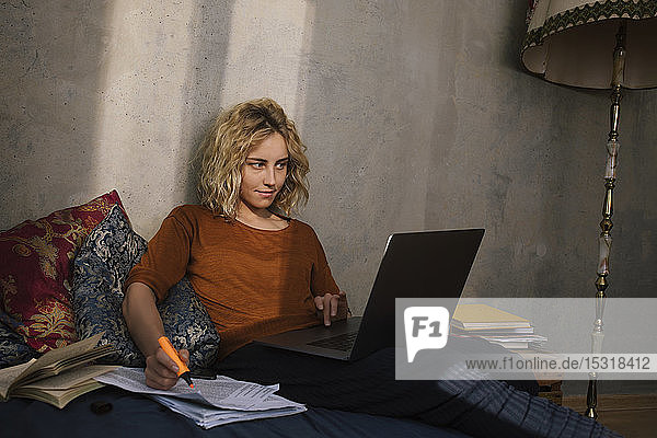 Porträt einer blonden Studentin  die am Bett sitzt und am Laptop arbeitet