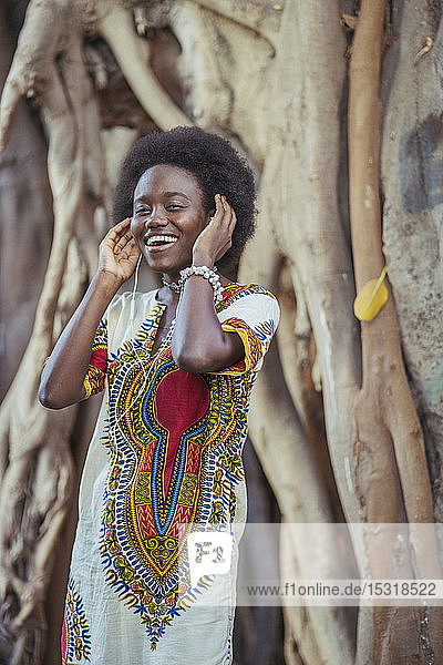 Lächelnde junge Frau steht mit Kopfhörern vor einem Baumstamm