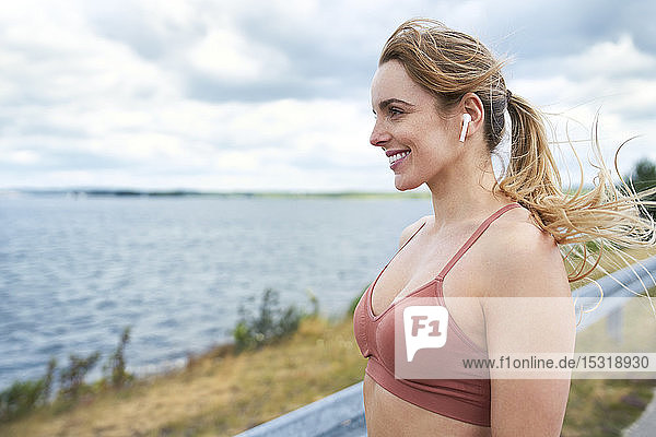 Porträt einer lächelnden sportlichen Frau mit drahtlosen Kopfhörern am Seeufer