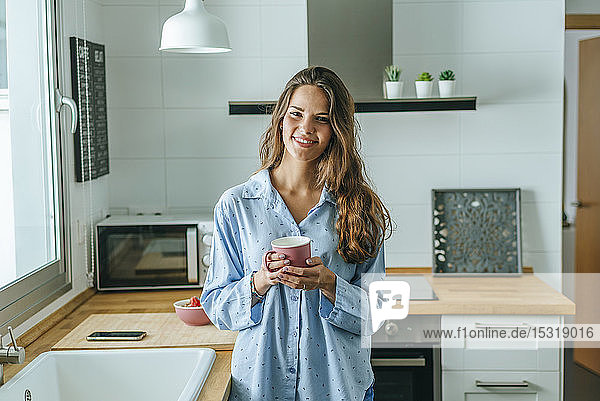 Porträt einer lächelnden jungen Frau mit Tasse Kaffee in Pyjama in der heimischen Küche