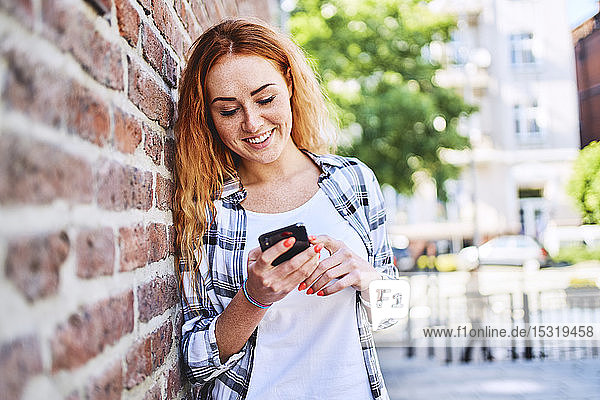 Young woman leaning against brick wall in the city while using smartphone