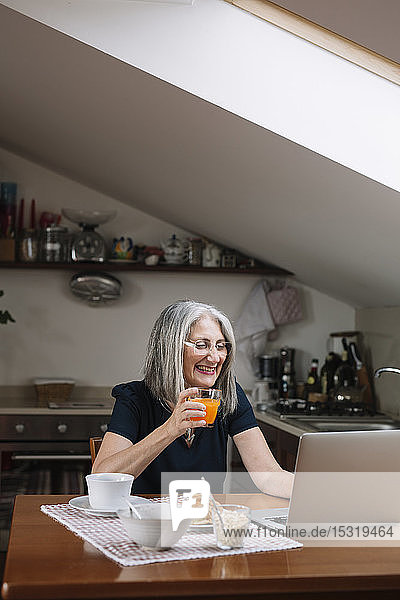 Porträt einer lachenden älteren Frau mit Laptop am Frühstückstisch