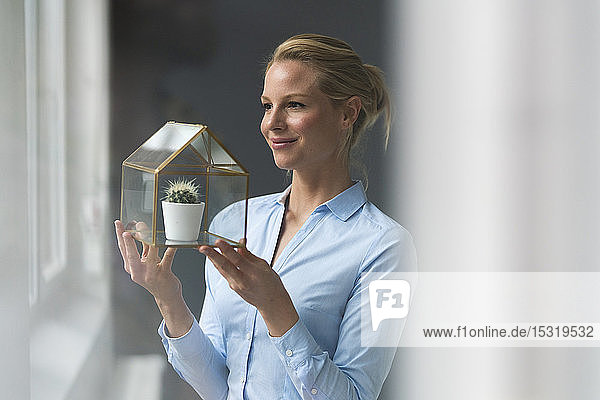 Lächelnde junge Geschäftsfrau hält Glashausmodell mit Kaktus in der Hand